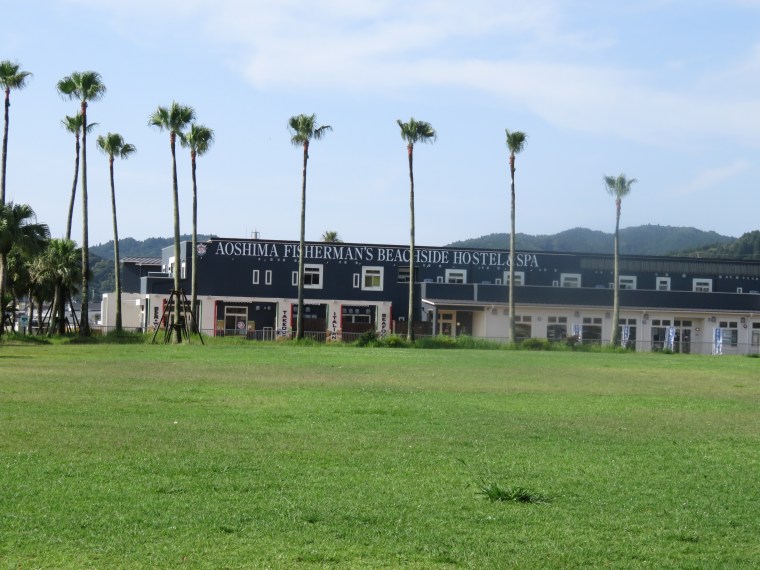 Aoshima Fisherman's Beachside Hostel & Spa building surrounded by palm trees and grass, with mountains in the background.