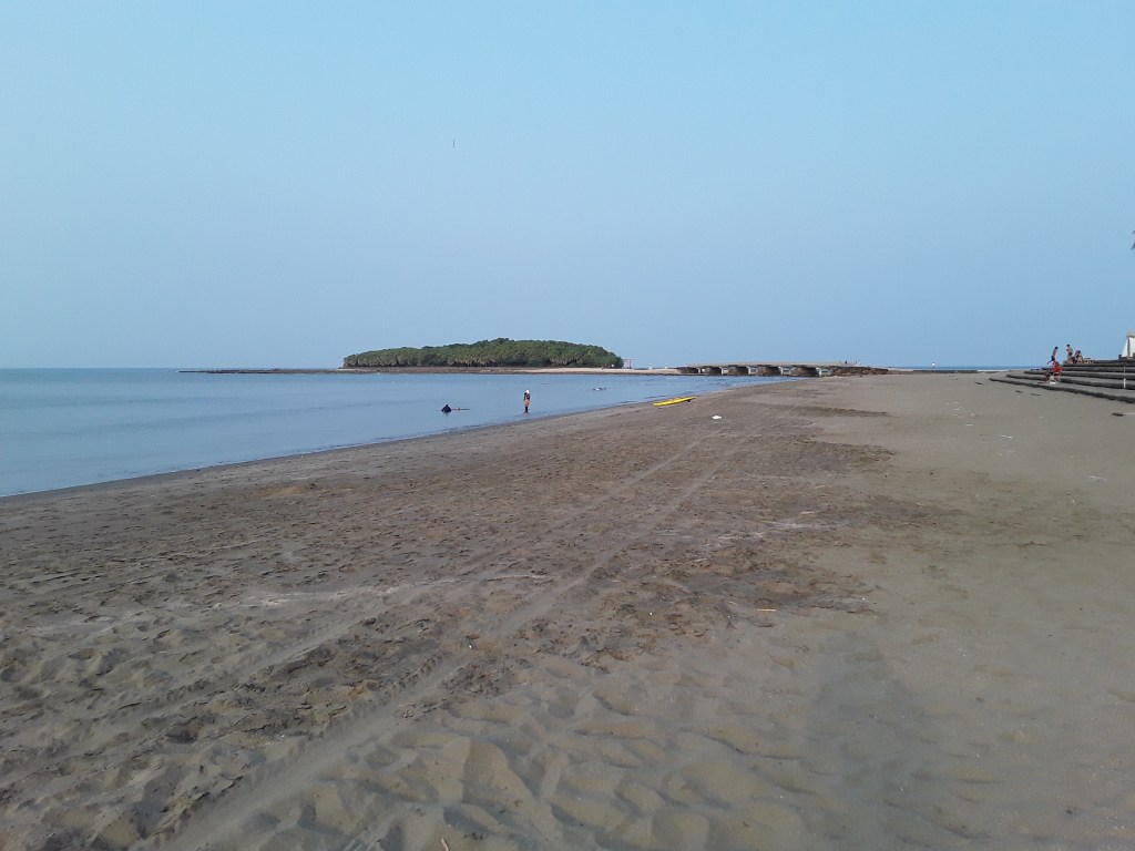A sandy beach with calm waters and a small island in the distance, featuring a bridge connecting the mainland.