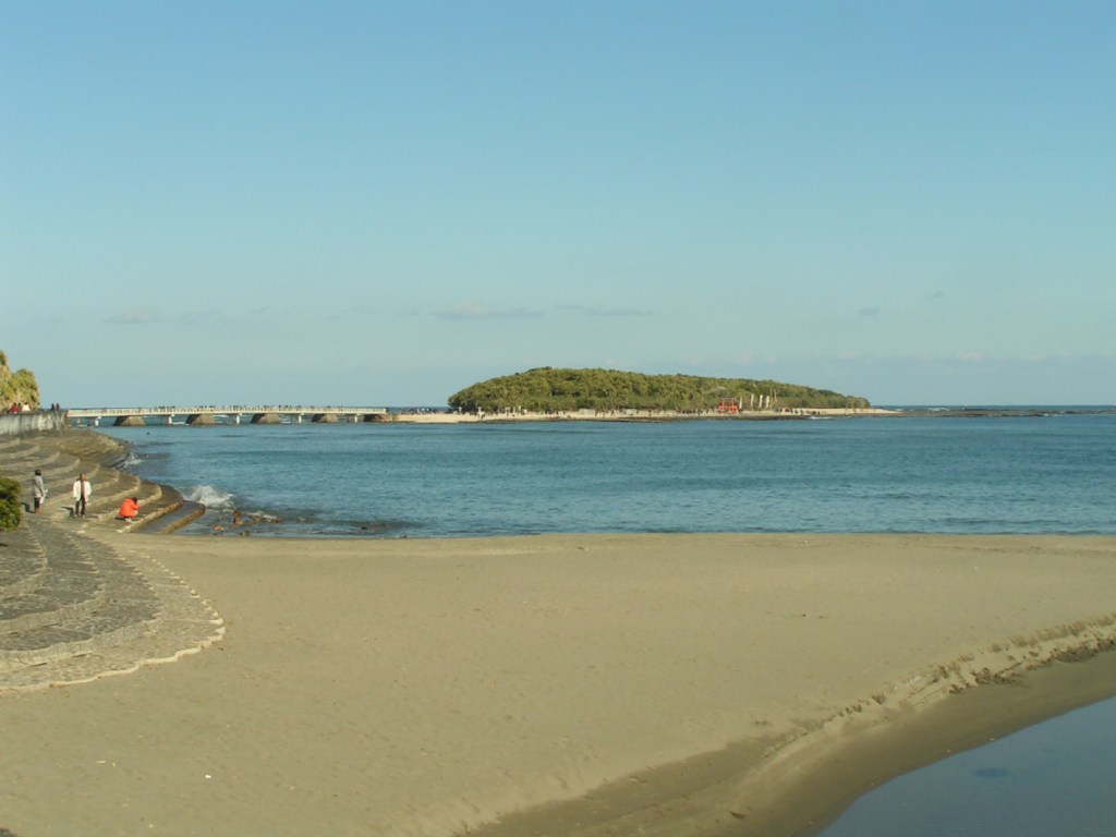 View of Aoshima Island connected to the mainland by a bridge, featuring a sandy beach and clear blue water.