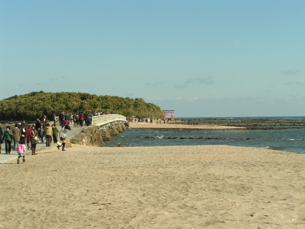 Aoshima Island connected to the mainland by a bridge with visitors walking along a sandy beach and rocky shoreline.
