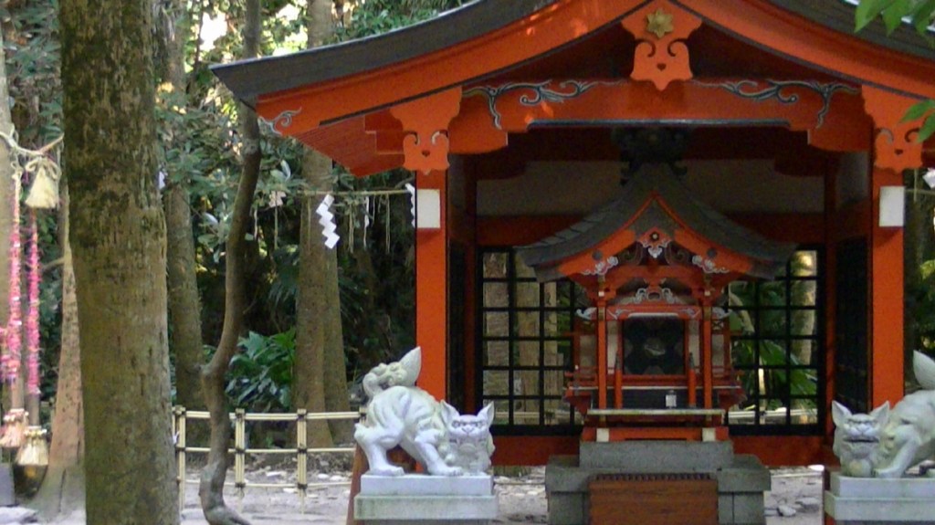 Aoshima Island shrine surrounded by lush trees, featuring a small red and black building with decorative roof and statues at the entrance.