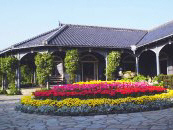 A historic building with a traditional Japanese roof surrounded by a colorful flower garden featuring various blooms in shades of pink, yellow, and blue.