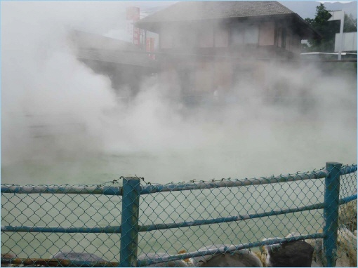 A steam-filled landscape near a hot spring with a fence in the foreground, showing a building partially obscured by mist.