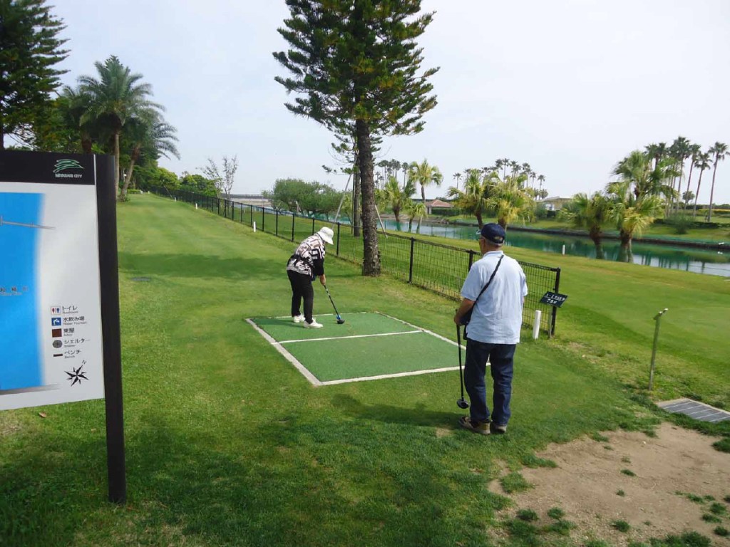 A scene of park golf being played in a lush green area with palm trees and water in the background. An individual is preparing to take a shot, while another person observes.