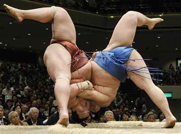 Two sumo wrestlers engaged in a match, demonstrating a close contest with one wrestler in a red mawashi and the other in a blue mawashi, surrounded by a crowd in a wrestling arena.