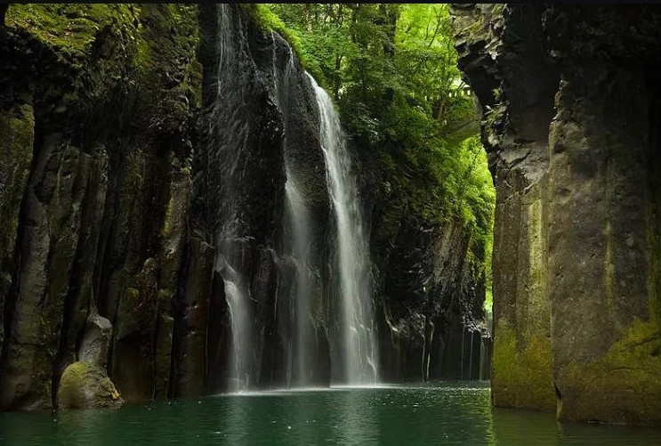 A scenic waterfall cascading down rocky cliffs into a tranquil pool, surrounded by lush greenery in Takachiho, Japan.