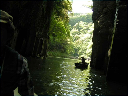 A serene river scene in Takachiho, Japan, framed by lush green cliffs and a person in a small boat paddling through the calm waters.