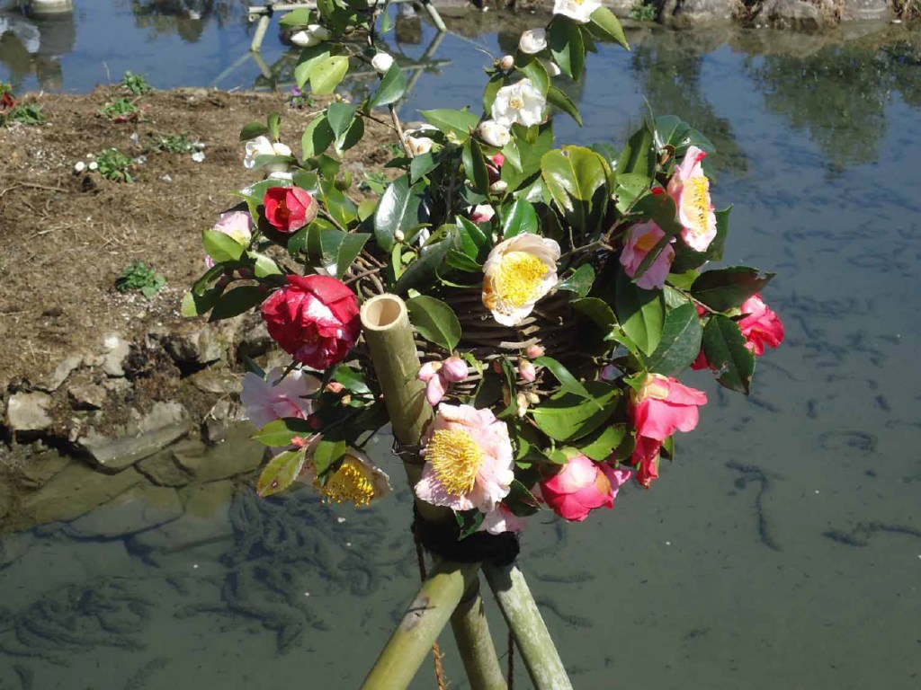 A decorative arrangement of camellia flowers in various colors, positioned on a bamboo stand beside a body of water.