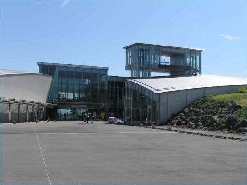 An exterior view of the Mount Unzen Disaster Memorial Hall featuring modern architecture, large glass windows, and a landscape of rocks and greenery.