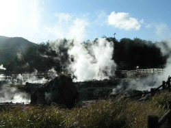 Steam rising from the geothermal area in Unzen, Japan, surrounded by hilly terrain and a wooden pathway.