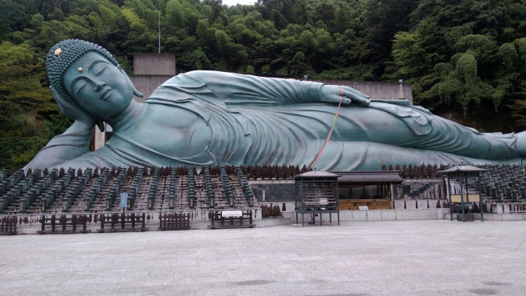 A large reclining Buddha statue surrounded by smaller statues, set in a serene outdoor environment with greenery in the background.