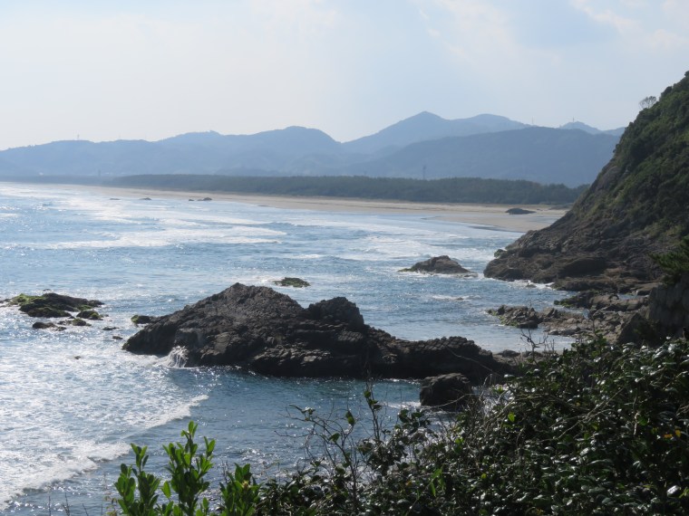 A scenic view of the Nichinan Coast in Miyazaki Prefecture, Japan, showcasing rocky shorelines, waves crashing onto the beach, and mountains in the background under a clear blue sky.