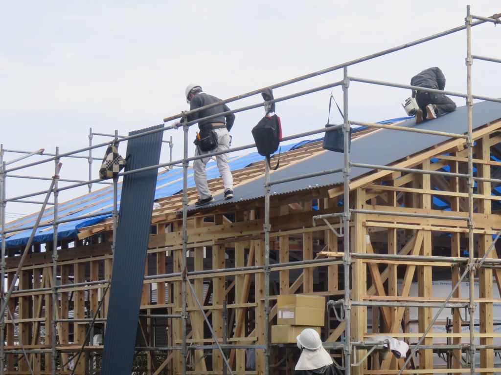 Workers installing roofing on a wooden house under construction in Japan, with scaffolding and building materials visible.