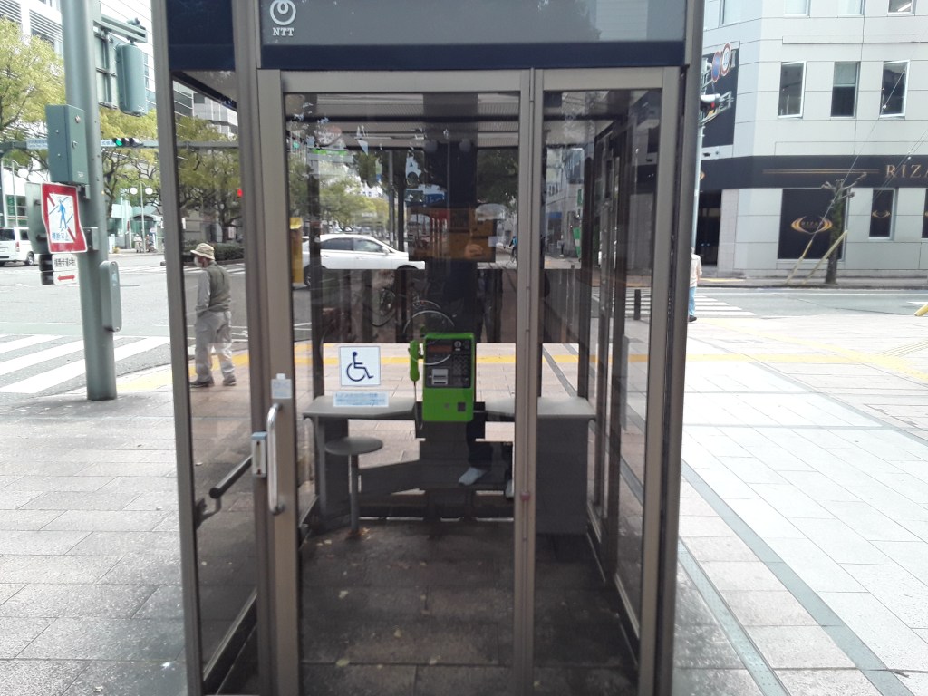 An empty phone booth in a city, featuring a wheelchair accessibility sign. The booth is glass-enclosed, with a bench inside and a green phone. There are pedestrians walking in the background.