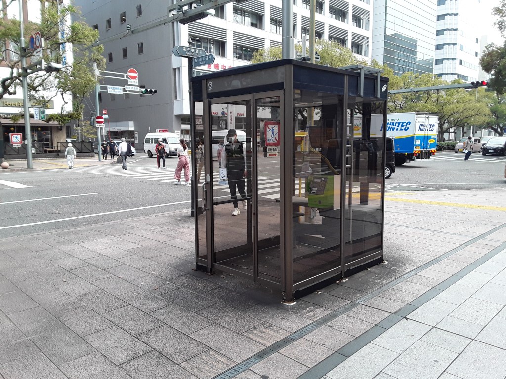 A modern telephone booth situated on a city sidewalk in Japan, with pedestrians crossing the street in the background.