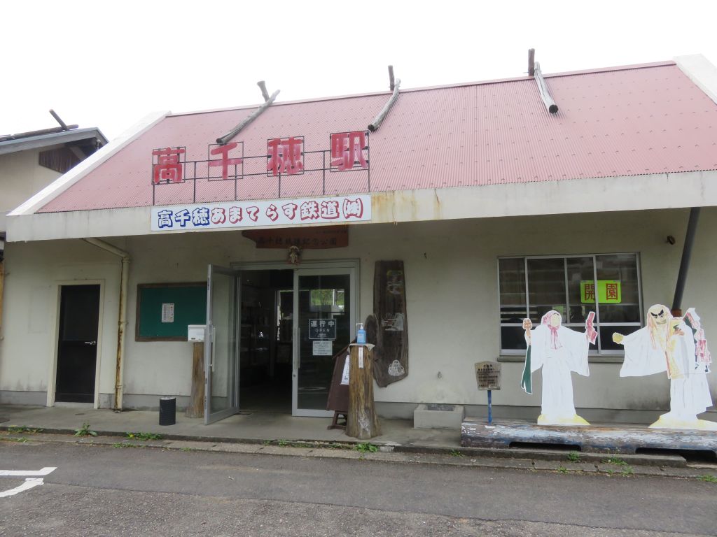 Entrance of a traditional Japanese building with a red roof and characters displayed prominently, surrounded by decorative cutouts of figures in traditional attire.