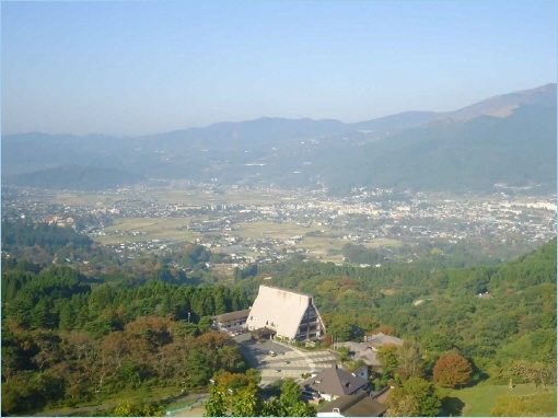 A scenic view of a valley in Kyushu, Japan, featuring a large building and surrounding mountains in the background.